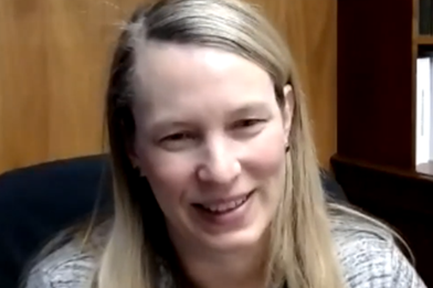 A smiling woman with long blonde hair appears on a video call from an office.