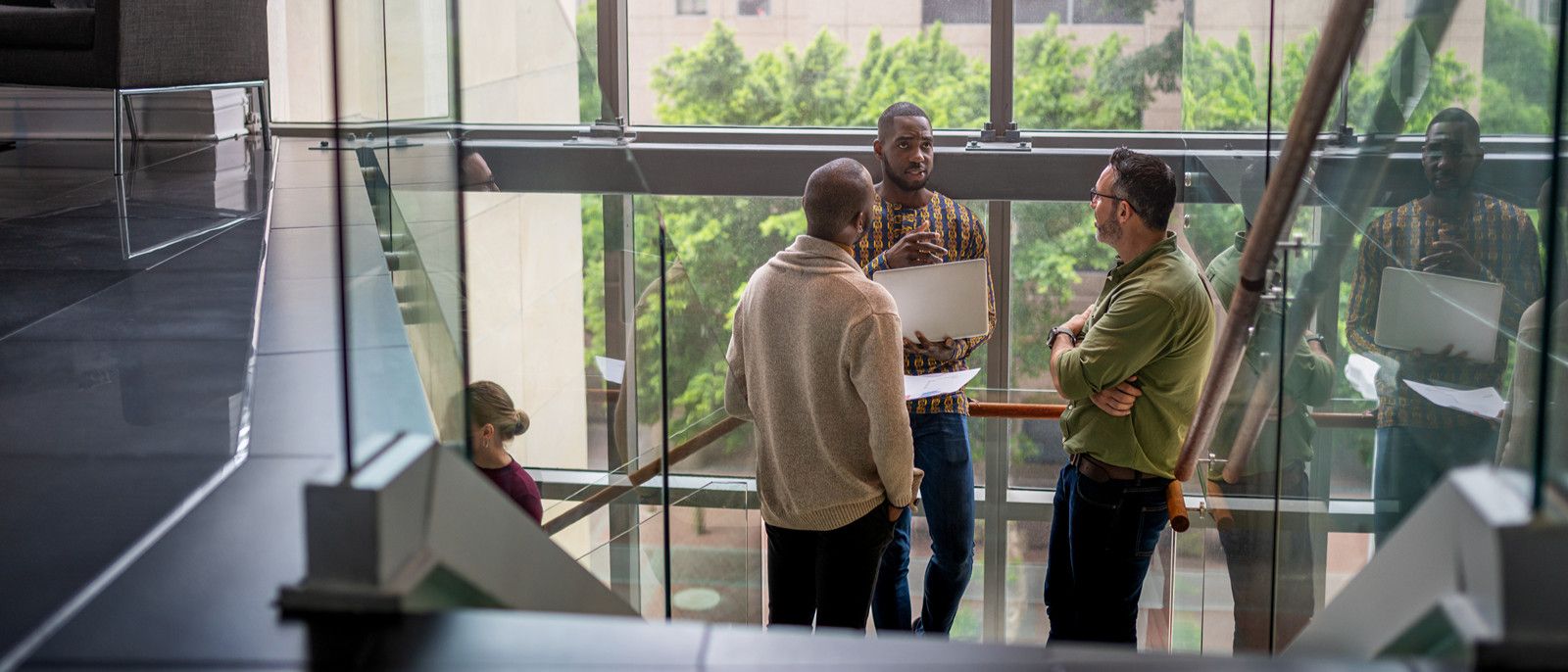People gather and talk on a sunlit stair landing inside a glass-fronted building.