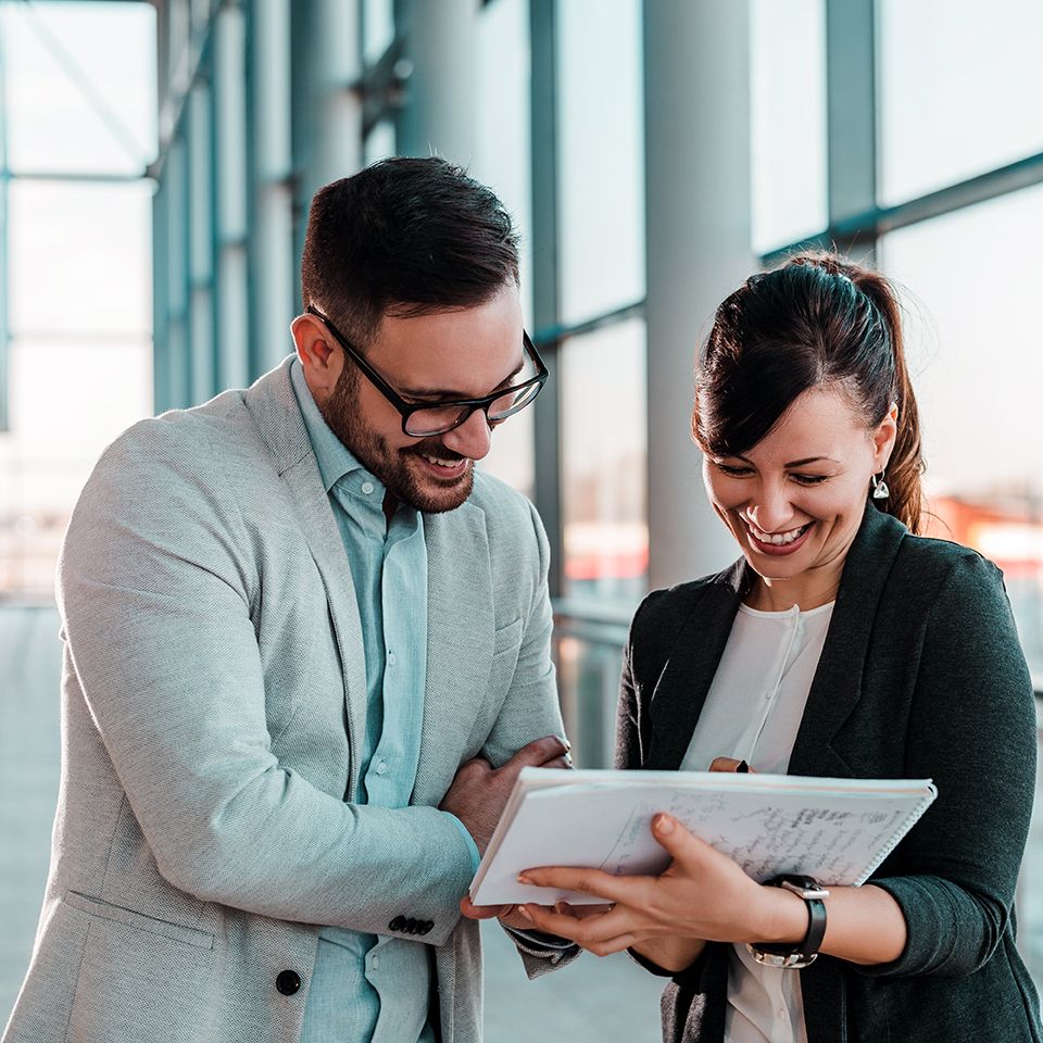 Two coworkers smile while reviewing a tablet beside large office windows.