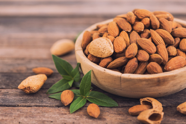 A wooden bowl filled with almonds beside loose nuts and green leaves.