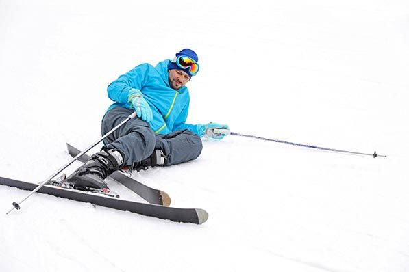 A skier in a blue jacket sits in the snow with skis crossed in front of them.