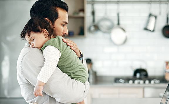 A man holds a sleeping young child against his shoulder in a kitchen.