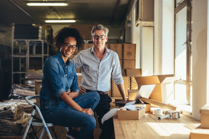 Two smiling people sit together in a workshop surrounded by tools and boxes.