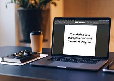 A laptop displays 'Completing Your Workplace Violence Prevention Program' beside a takeaway cup and notebook.