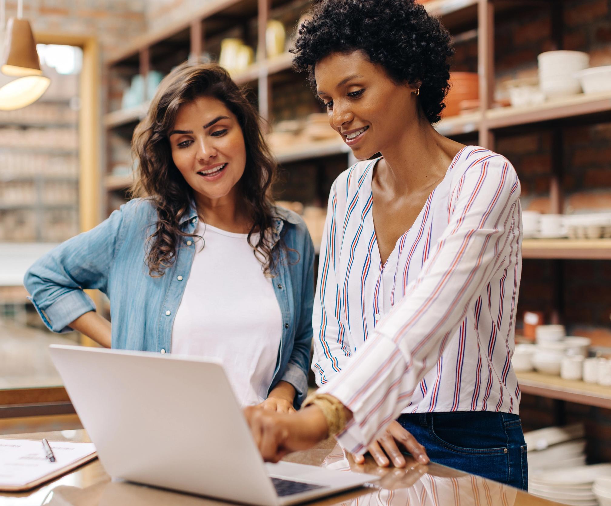Two women work together on a laptop in a shop lined with pottery.