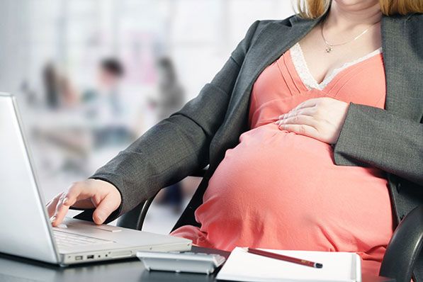A pregnant person rests a hand on their belly while working at a laptop.
