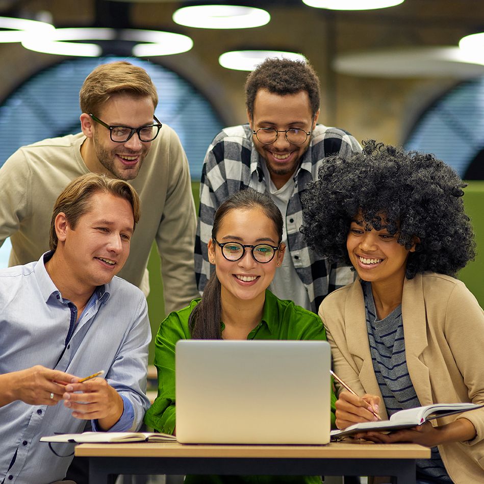 Five coworkers gather around a laptop, smiling at its screen.