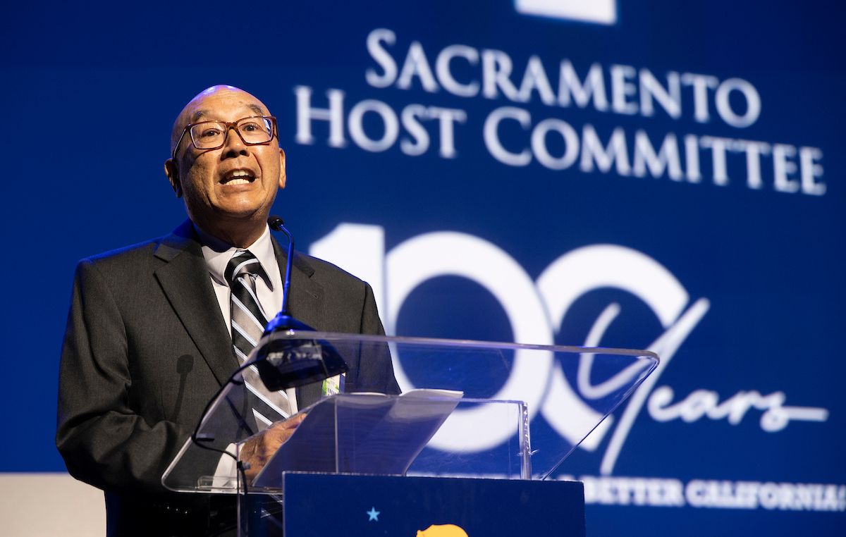 An older East Asian man in a suit and glasses speaks at a podium in front of a blue screen that says "Sacramento Host Committee 100 Years".
