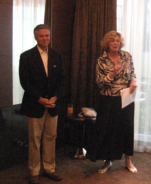 A man and woman stand before curtains in a meeting room, with the woman holding papers.