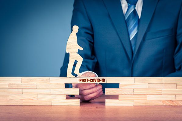 A suited person places a Post-COVID-19 block across a gap beneath a wooden walking figure.
