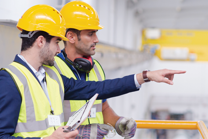 Two workers wearing yellow hard hats and reflective vests review a work area, with one pointing.
