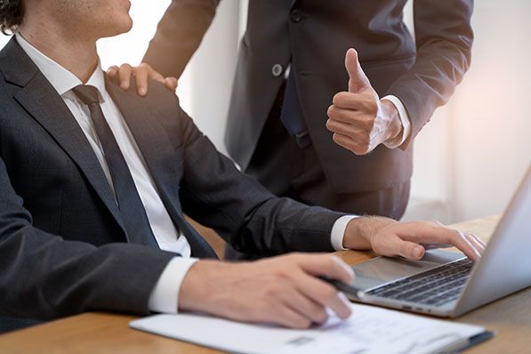 A standing colleague gives a thumbs-up and rests a hand on a seated laptop user's shoulder.