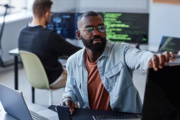 An office worker adjusts a monitor beside a laptop as a colleague works at screens displaying code.