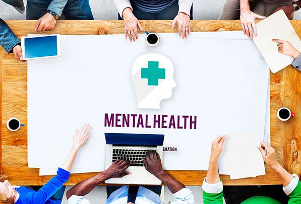 People sit around a table with Mental Health lettering and a head silhouette containing a medical cross.
