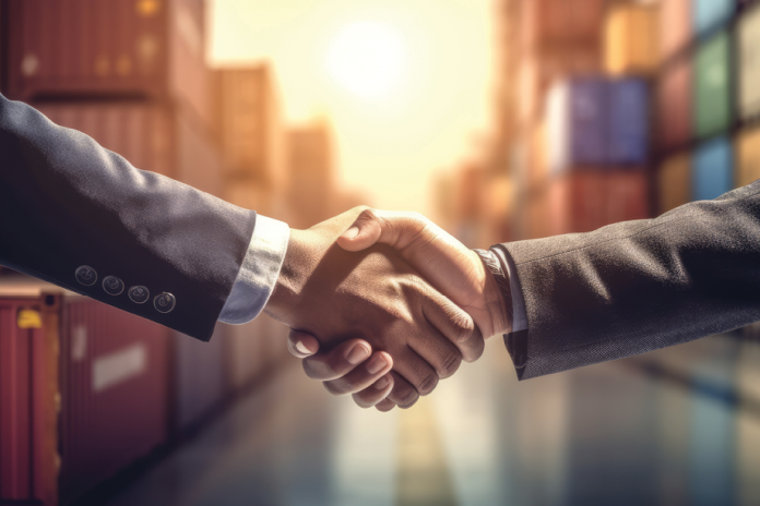 Two suited people shake hands in front of shipping containers at sunset.