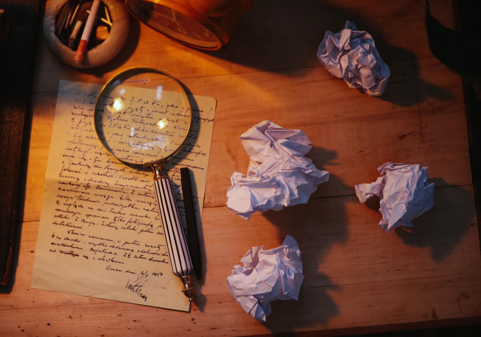 A handwritten letter under a magnifying glass in lamplight, surrounded by crumpled sheets of paper