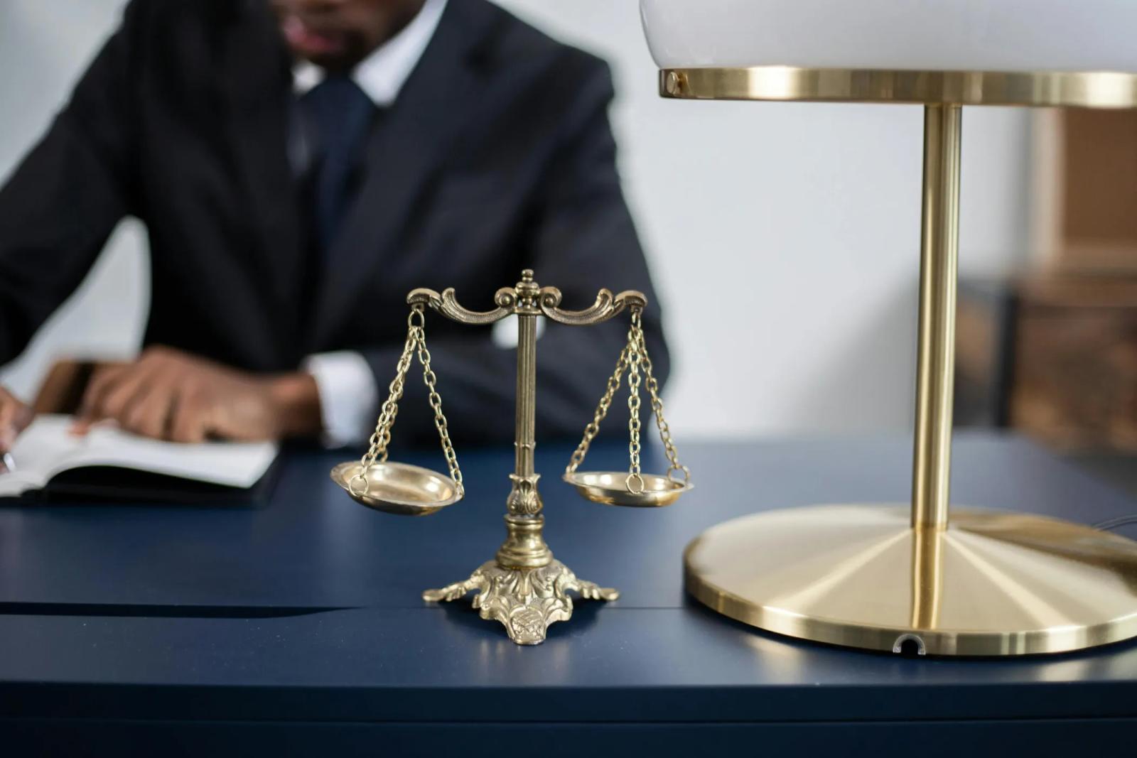 Brass scales of justice on a desk, with a person in a suit reading behind them