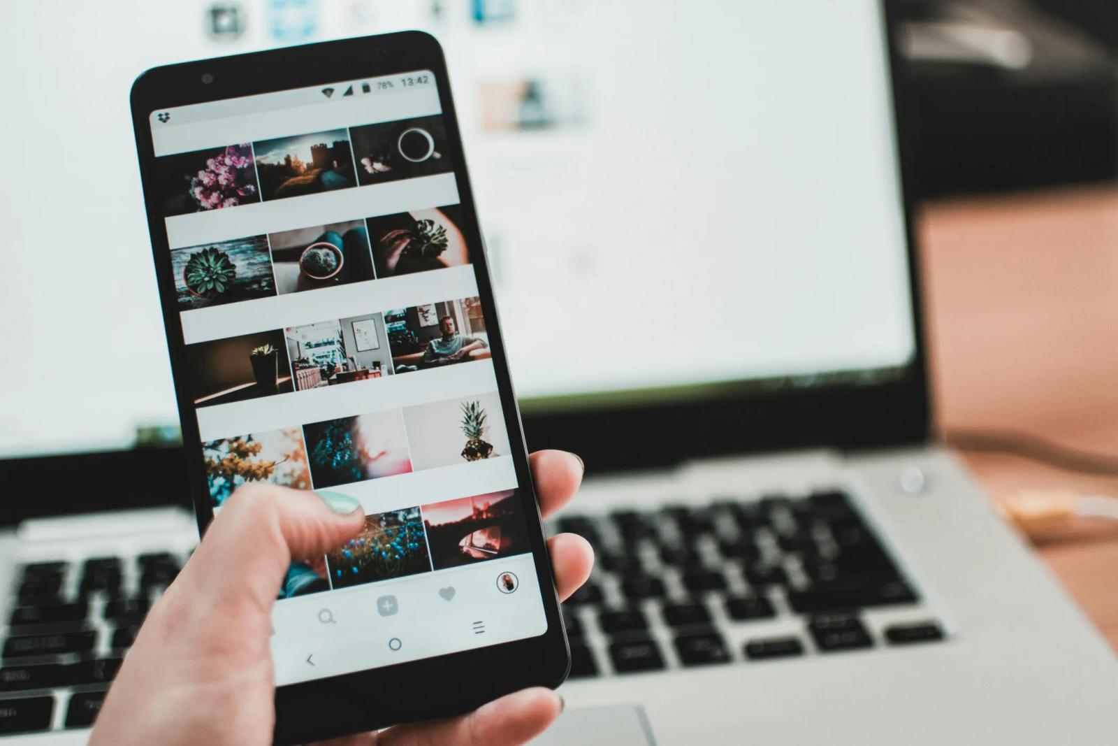 A hand holding a phone open on a grid of photos, a laptop screen behind it