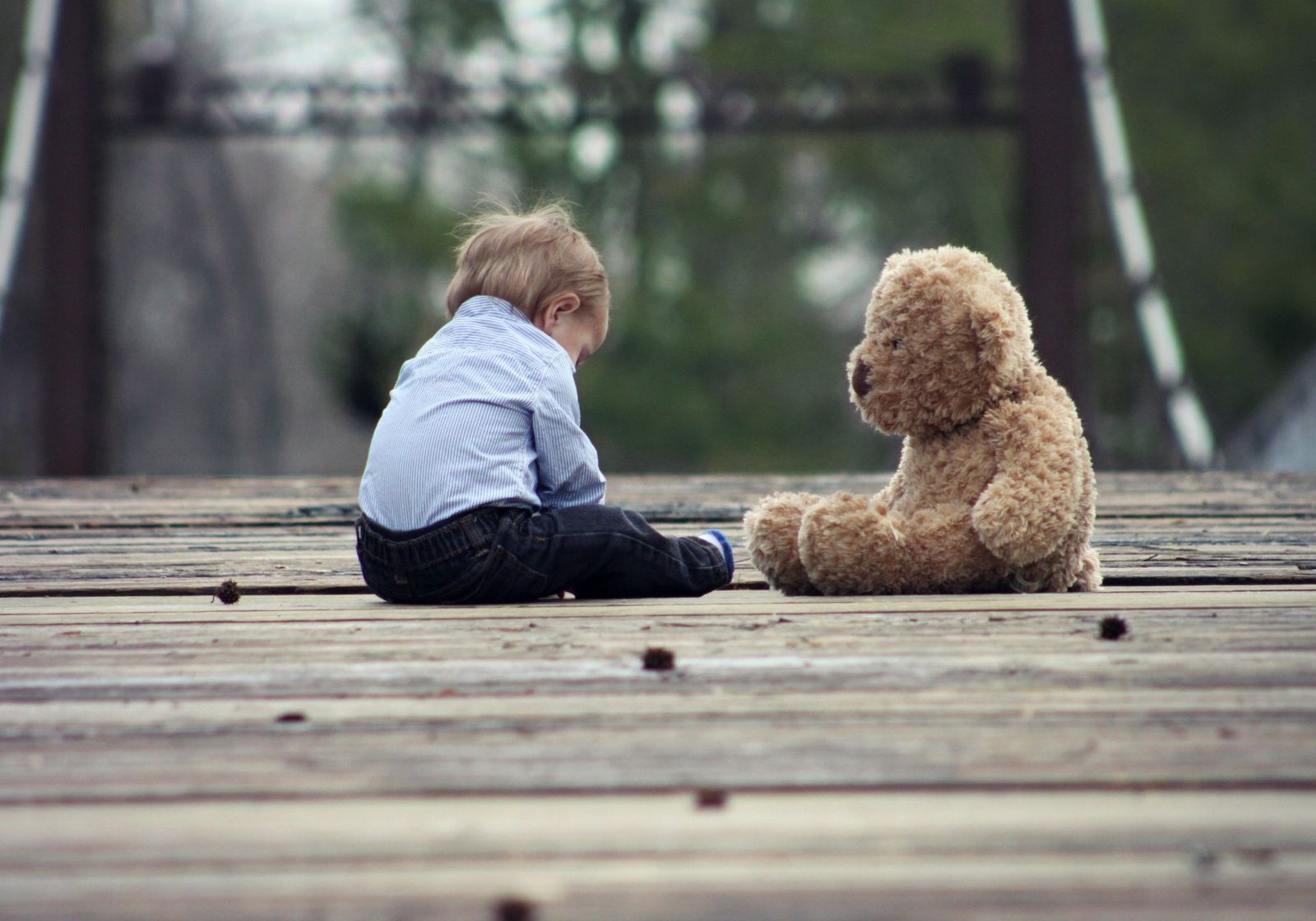 A toddler sitting alone on a wooden bridge, facing a teddy bear