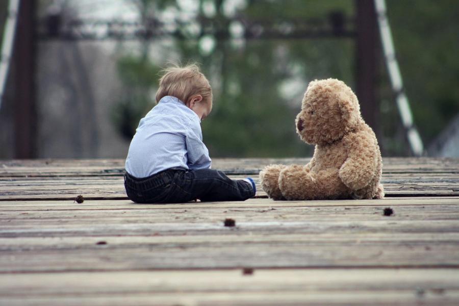 A toddler sitting alone on a wooden bridge, facing a teddy bear