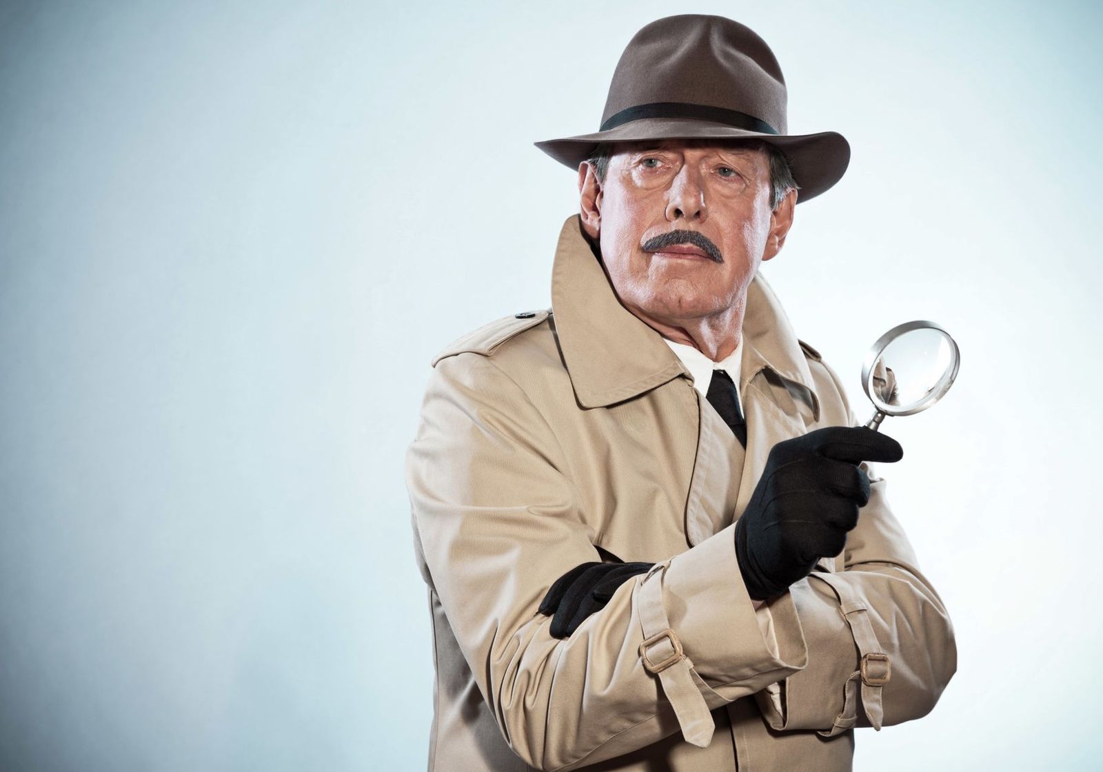 A man in a fedora and trench coat holding up a magnifying glass