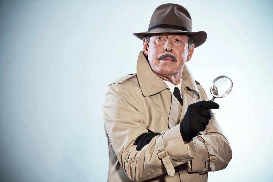A man in a fedora and trench coat holding up a magnifying glass