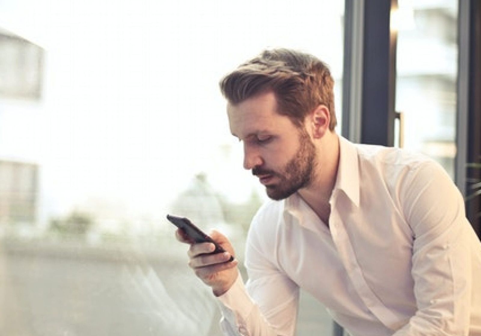 A man standing at a window, studying the screen of his phone
