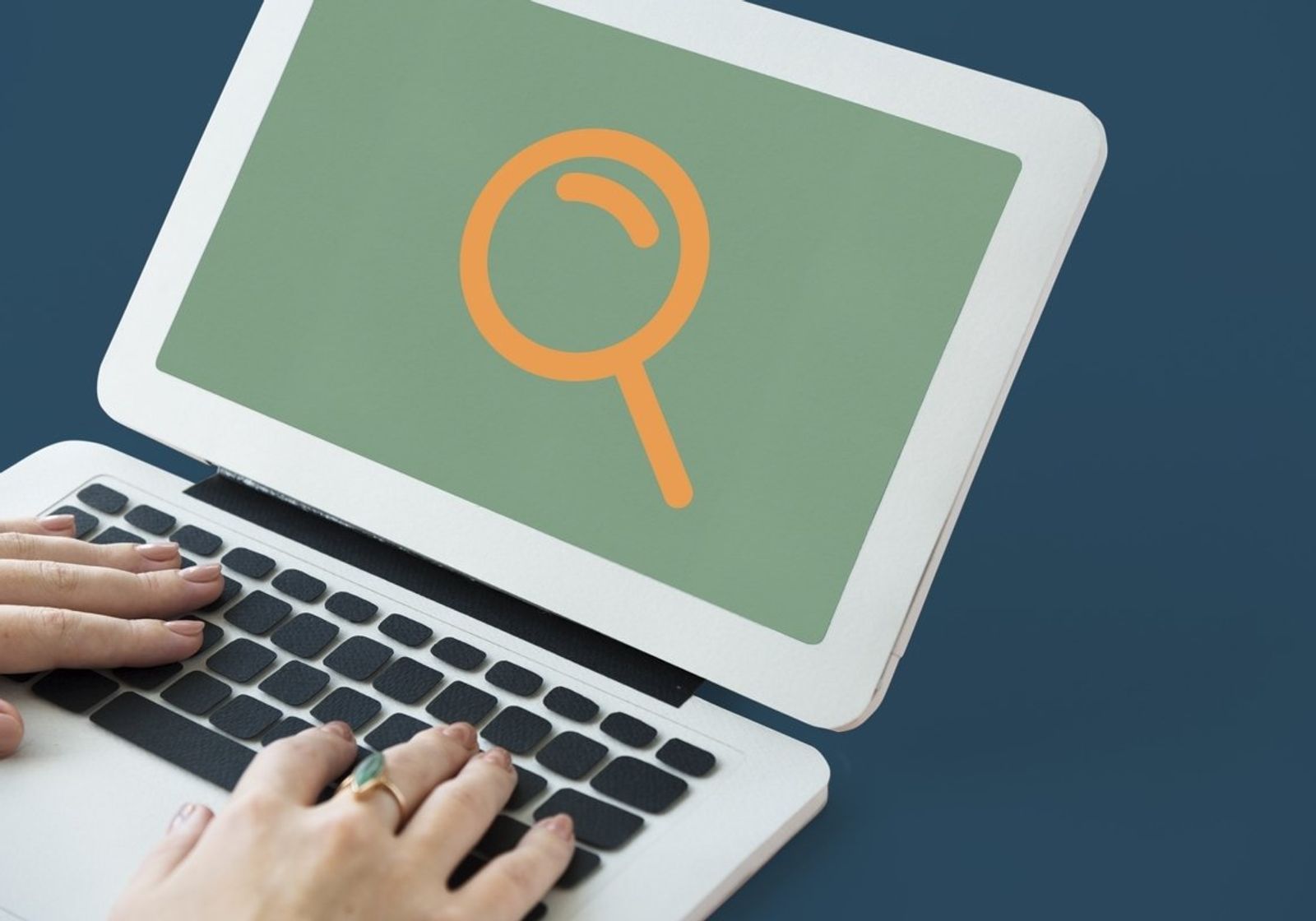 Hands typing on a laptop whose screen shows a large magnifying glass icon