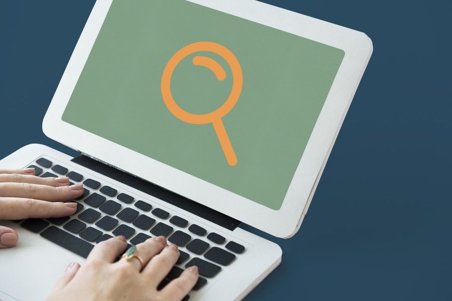 Hands typing on a laptop whose screen shows a large magnifying glass icon