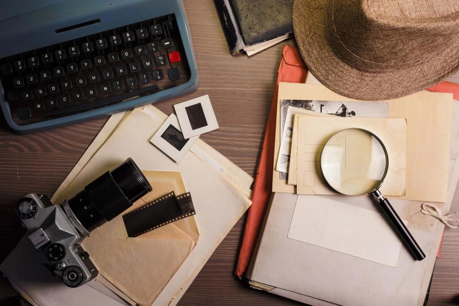 A flat lay of vintage detective gear: a typewriter, a film camera, a magnifying glass and a fedora