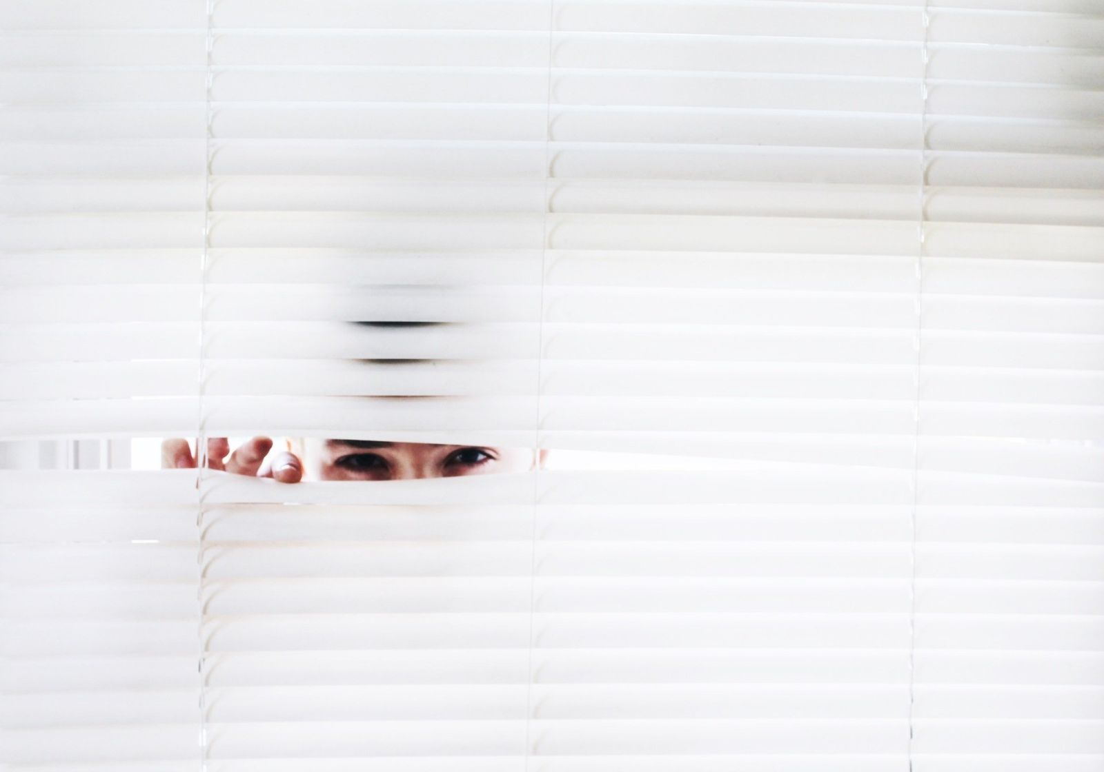 A pair of eyes peering out between the slats of a white venetian blind