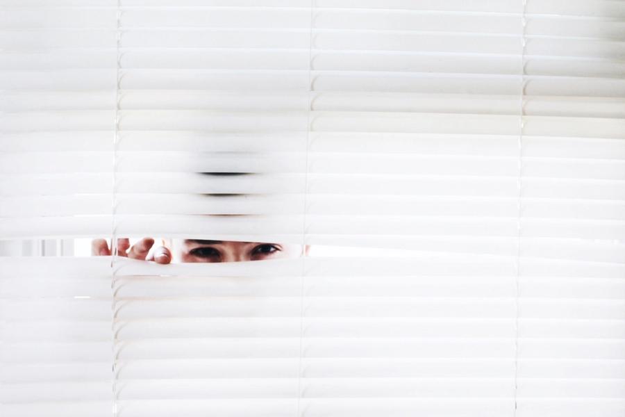 A pair of eyes peering out between the slats of a white venetian blind
