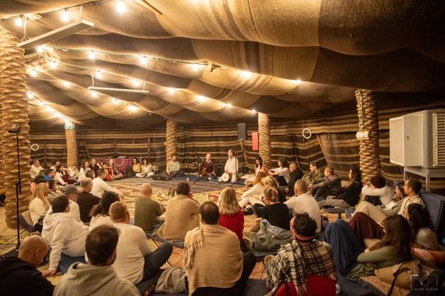 A large group sitting in a circle in a tent hall lit with string lights