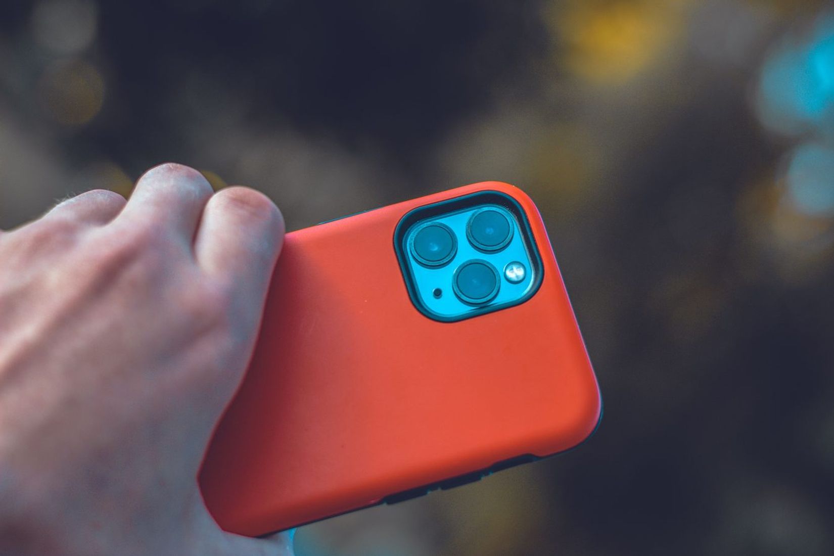 A close-up view of a hand holding a smartphone with an orange case and three camera lenses.