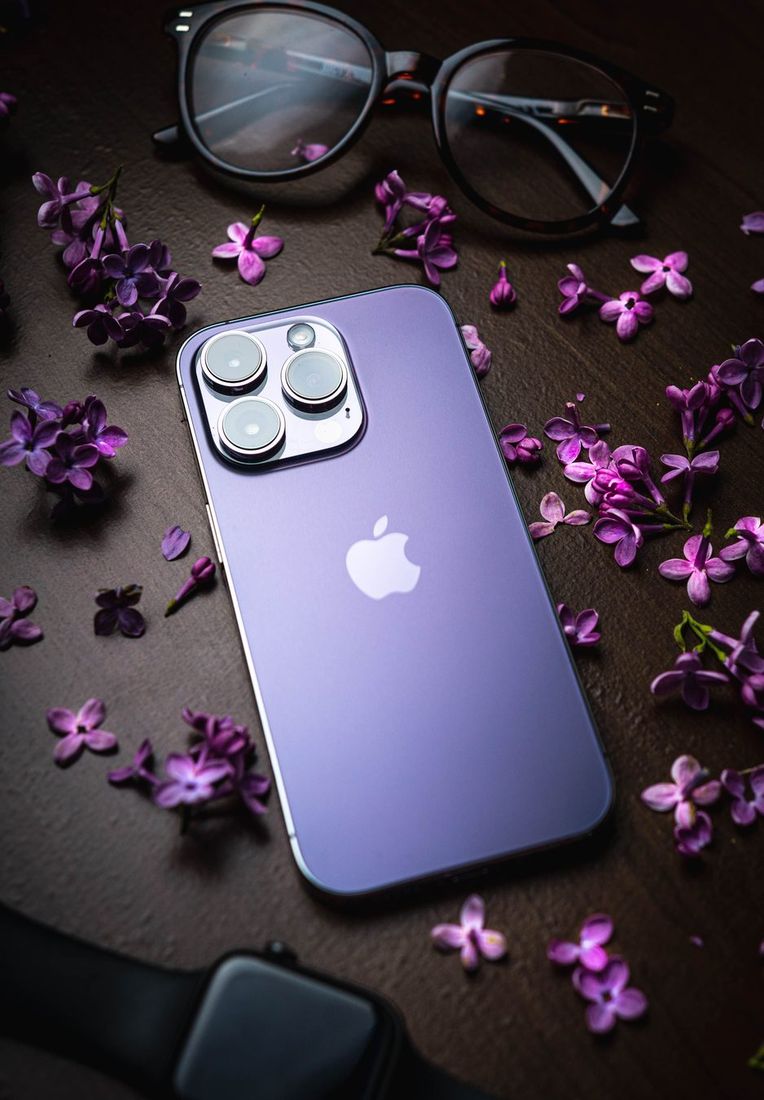 Stylish purple smartphone surrounded by lilac flowers and eyeglasses on a dark wooden table.
