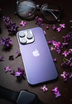 Stylish purple smartphone surrounded by lilac flowers and eyeglasses on a dark wooden table.