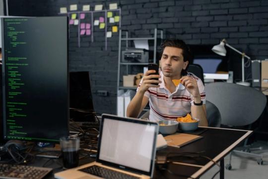 Programuotojas žiūri į telefoną prie biuro stalo