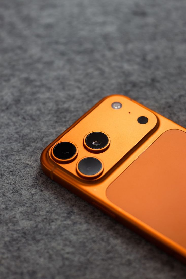 Oranžinis išmanusis telefonas su triguba kamera ant švelnaus pilko audinio