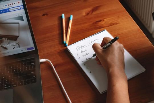 Person writing notes next to a laptop in a home workspace.