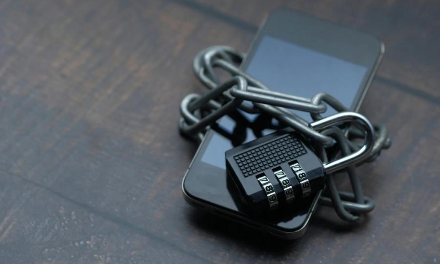 Iš arti nufotografuotas išmanusis telefonas, apsivyniojęs grandine su spyna, simbolizuojantis stiprią saugą