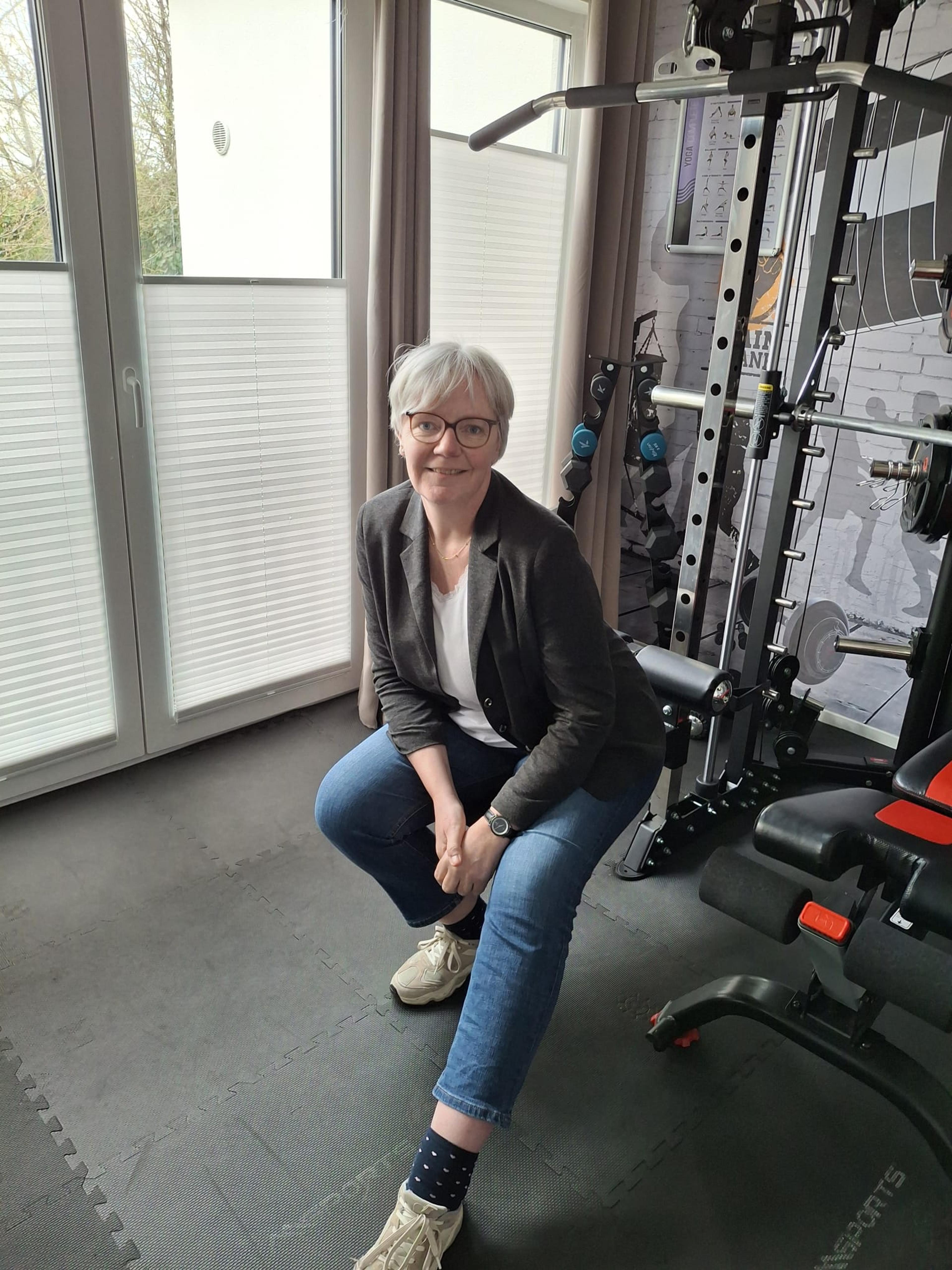 Ältere Frau mit Brille in Freizeitkleidung sitzt in Heimfitnessraum vor Fenster. Neben ihr ein Fitnessgerät.