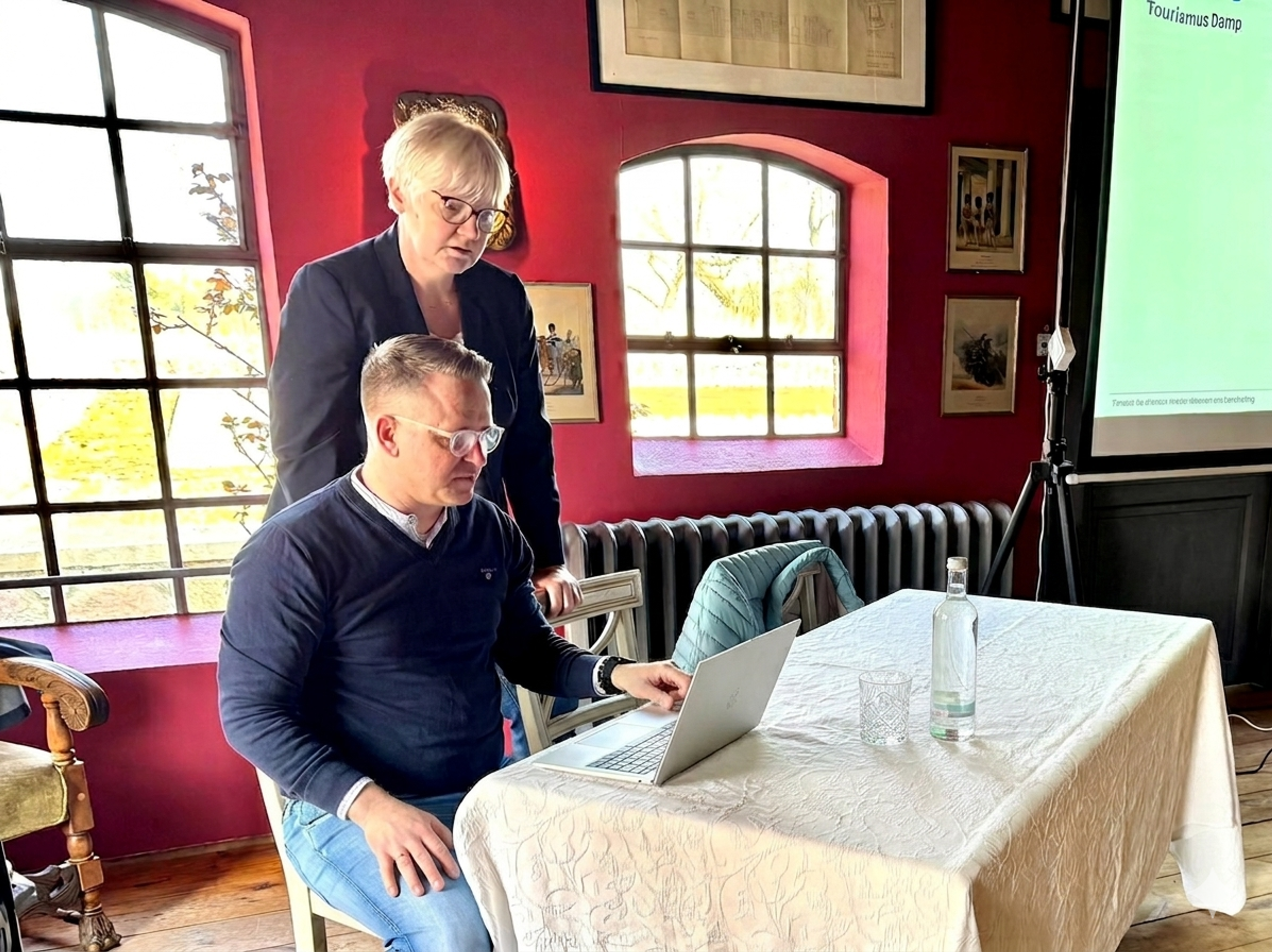 Ricci und Sebastian arbeiten gemeinsam an einem Laptop an einem runden Tisch, umgeben von roten Wänden und großen Fenstern.