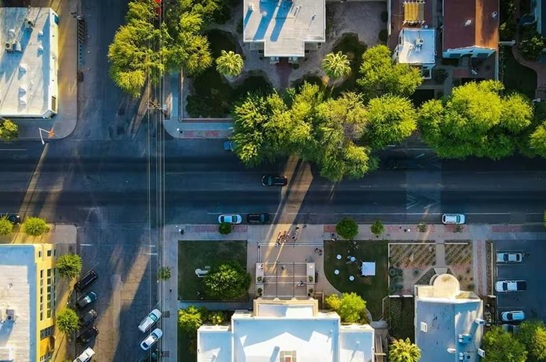 Overhead view of a city street with buildings, green trees, and parked cars under late afternoon sun.
