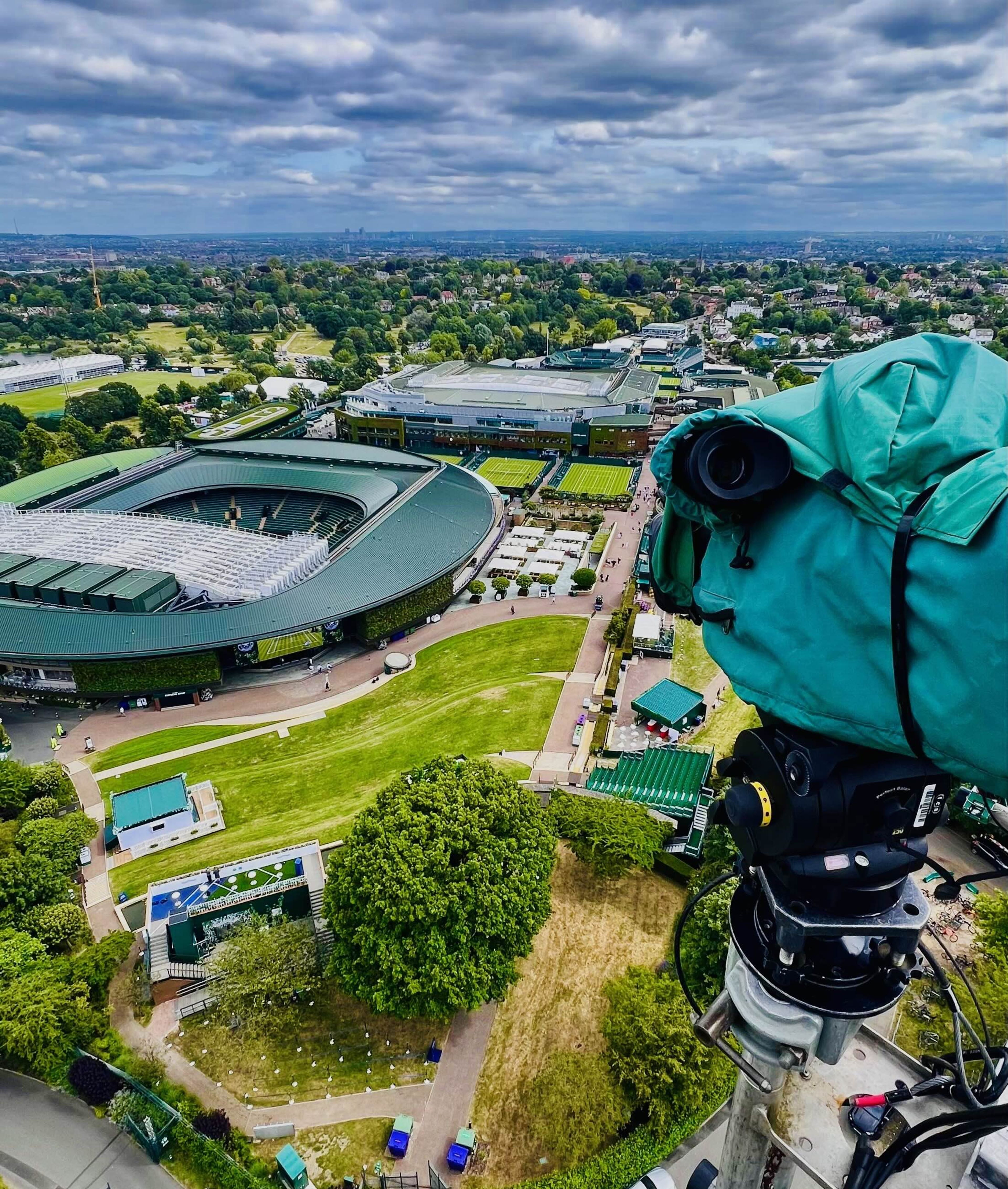 A broadcast camera under a green rain cover, mounted high above the Wimbledon grounds and looking down on the show courts.