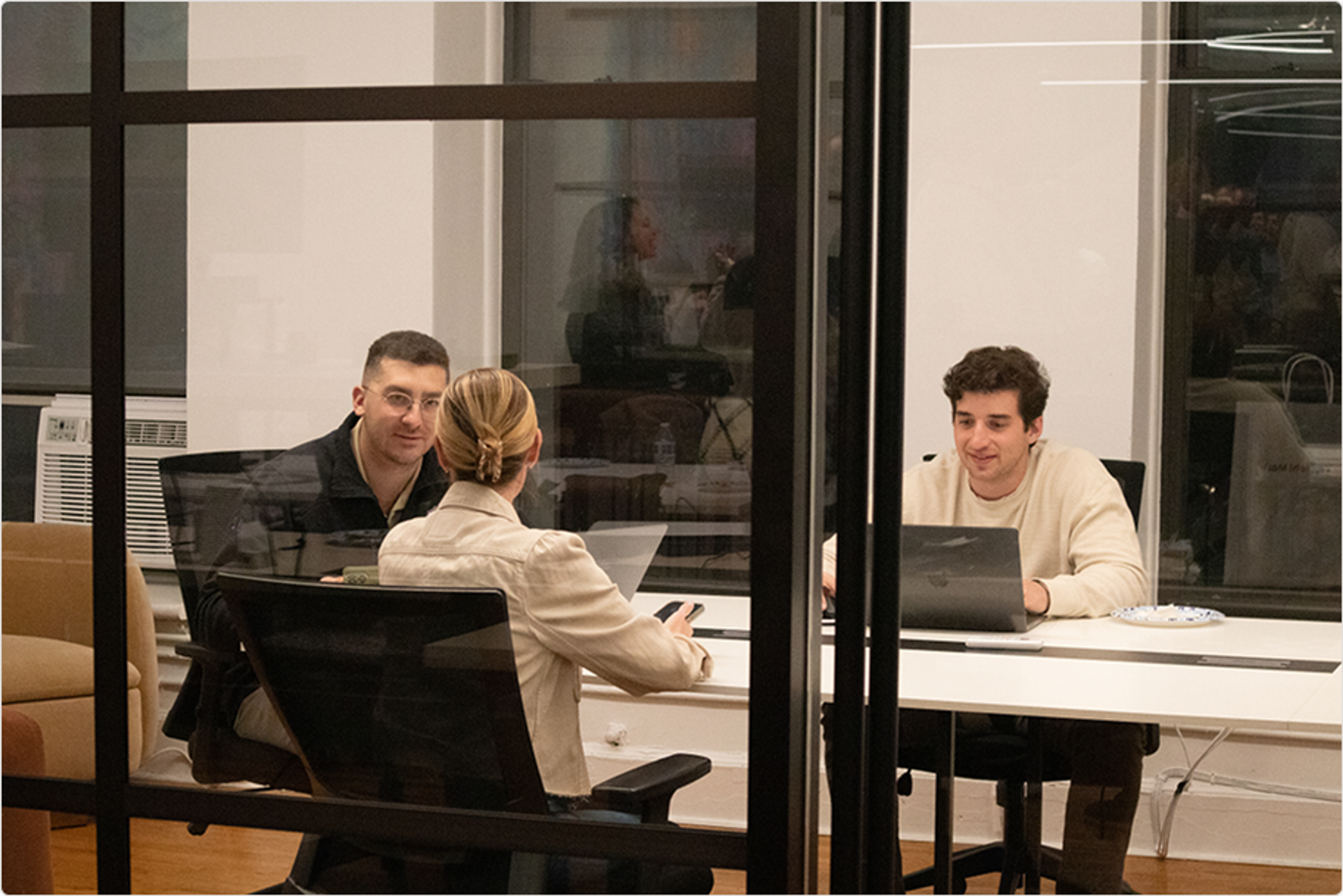 Three people in an office: two conversing and one working on a laptop, viewed through a glass wall.