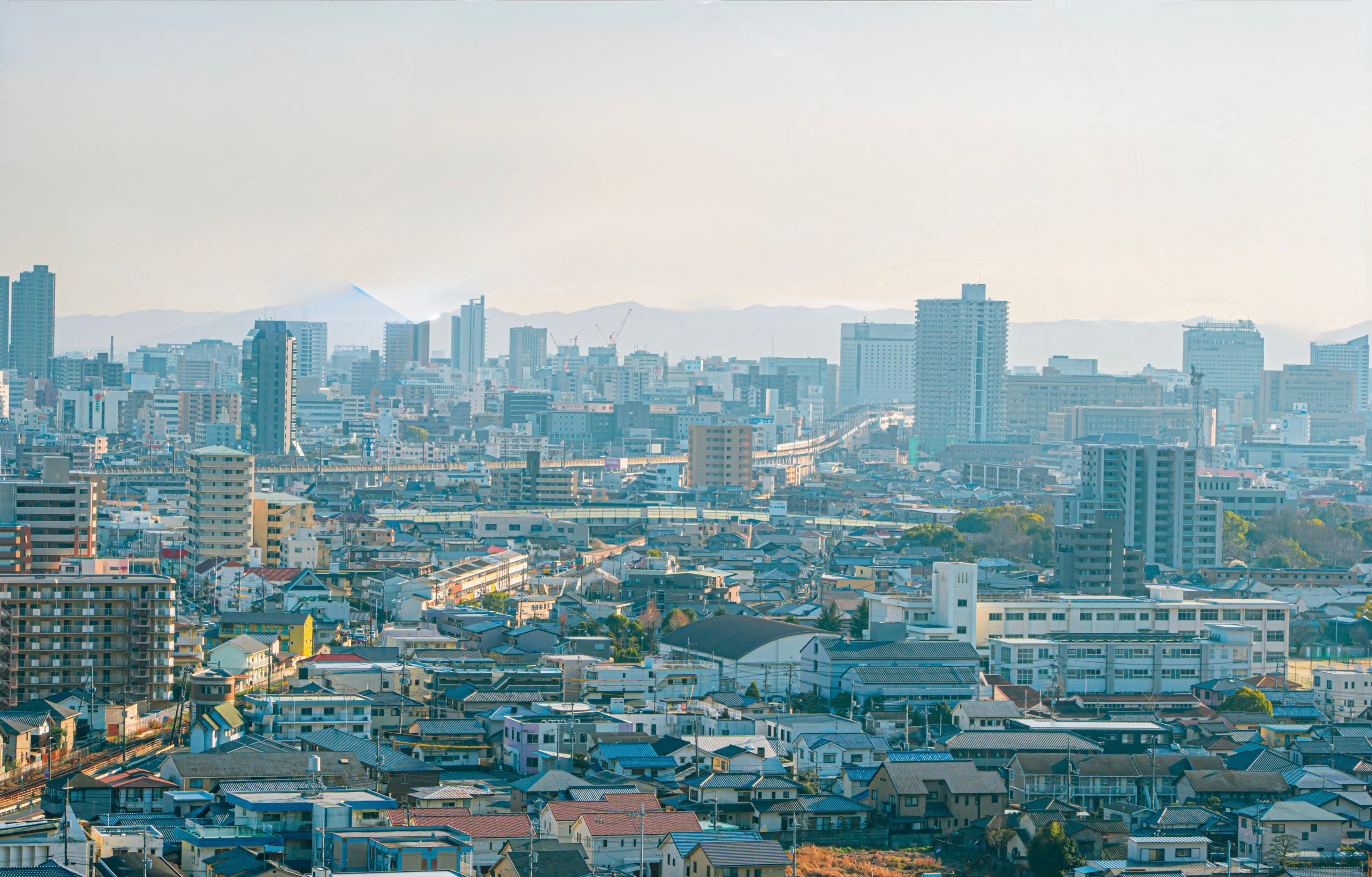The skyline of Okayama, low rooftops in the foreground giving way to high-rise towers and mountains beyond.