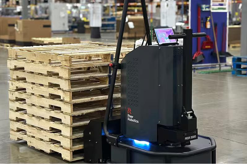 A gray robotic pallet mover stands next to a tall stack of wooden pallets in a warehouse.