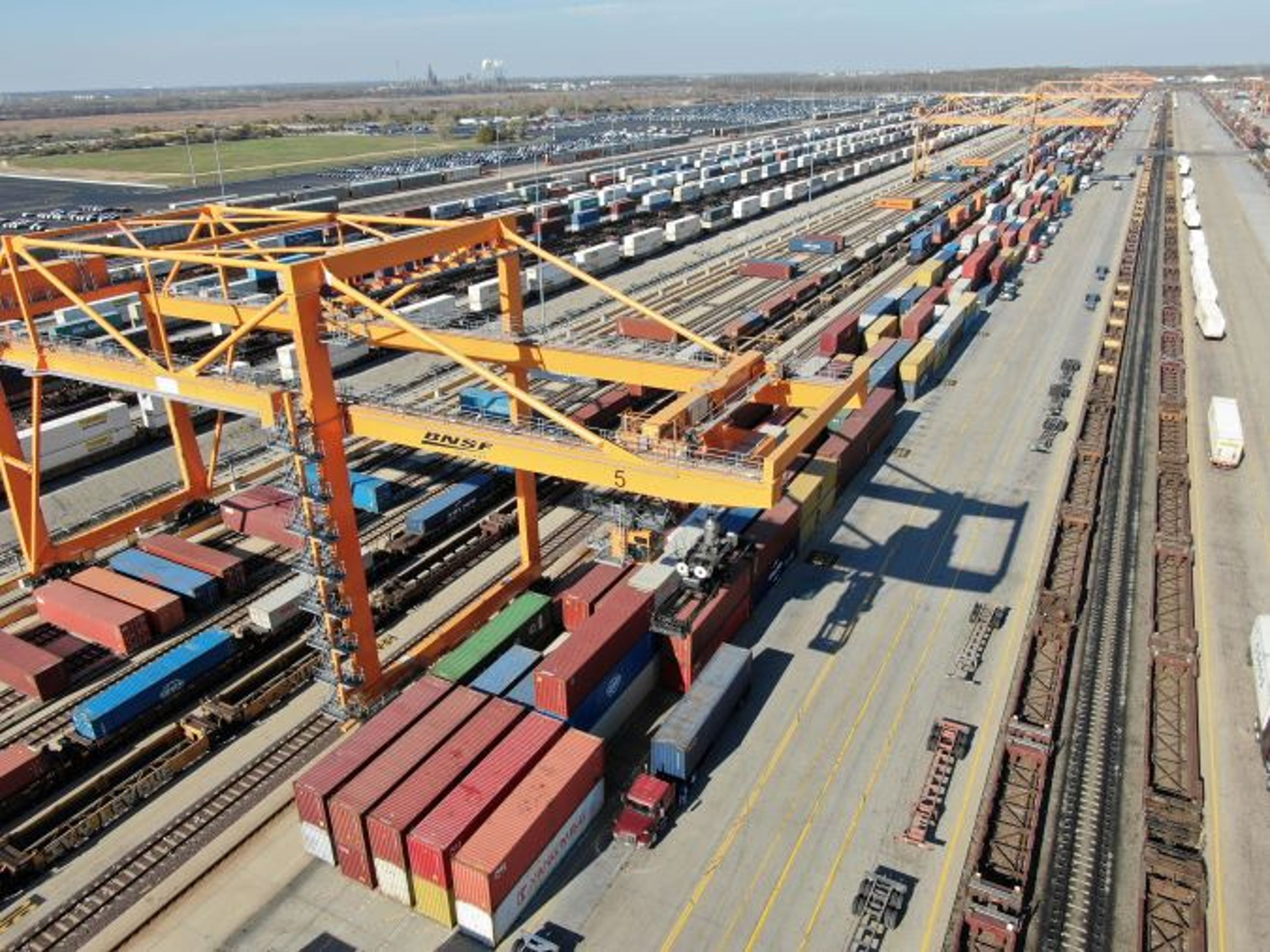 Aerial view of a BNSF intermodal facility, a yellow gantry crane straddling container stacks beside parallel tracks of freight cars.
