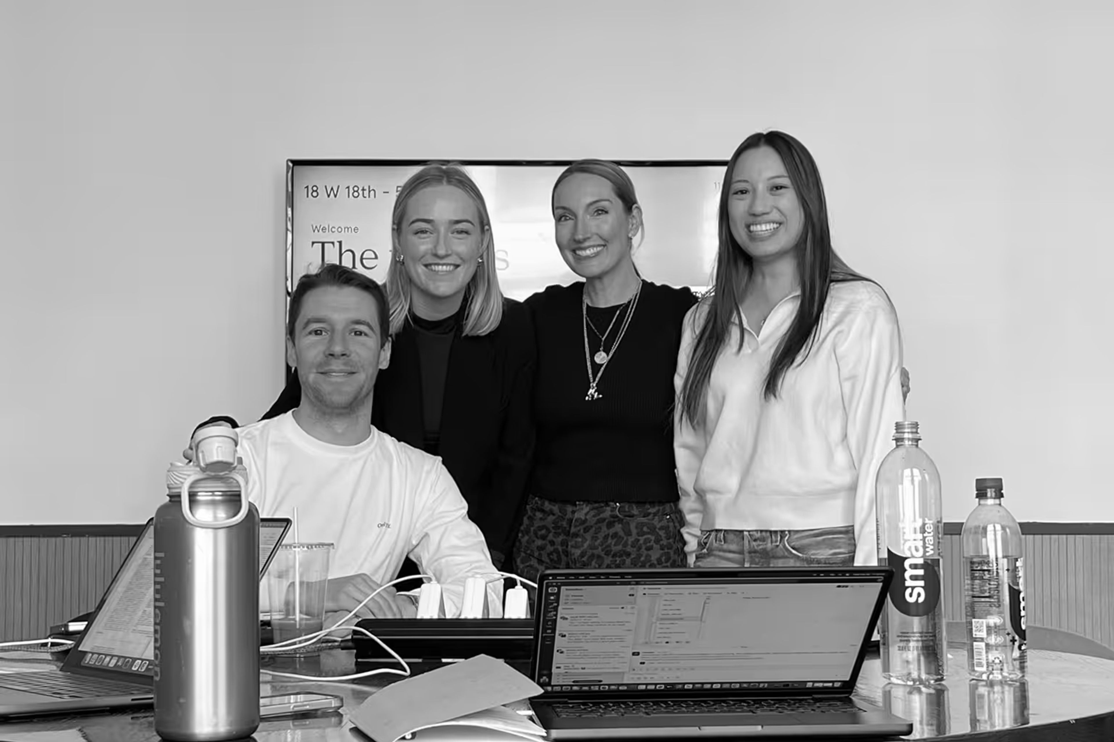Four smiling people, three women and one man, in an office setting with laptops and water bottles on a table.
