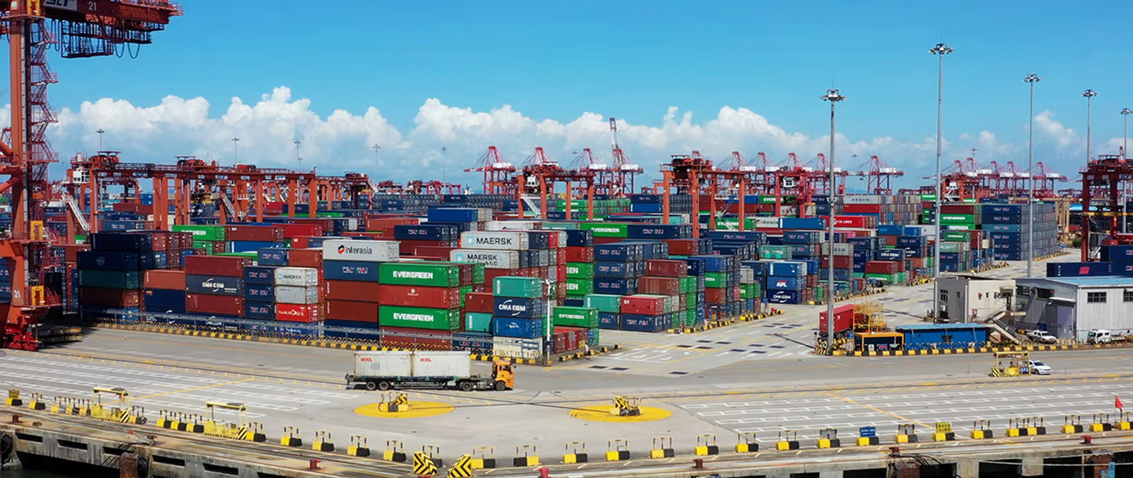 A vast container port with stacks of colorful shipping containers and numerous red gantry cranes under a blue sky.